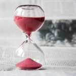 Close-up of a transparent hourglass with pink sand flowing, placed on a newspaper background.